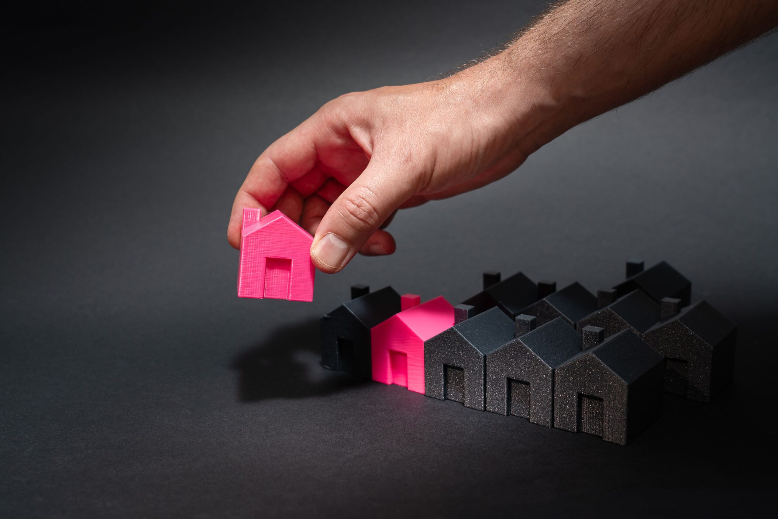 A hand selects a pink mini house among black models, symbolizing unique choice.